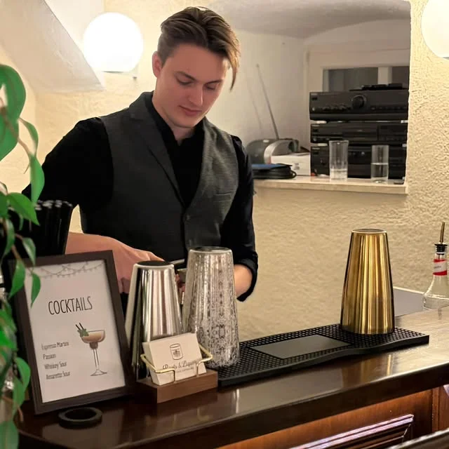 Ein Roots & Liquor Barkeeper in schwarzem Hemd und grauer Weste bereitet Drinks an der Bar zu.